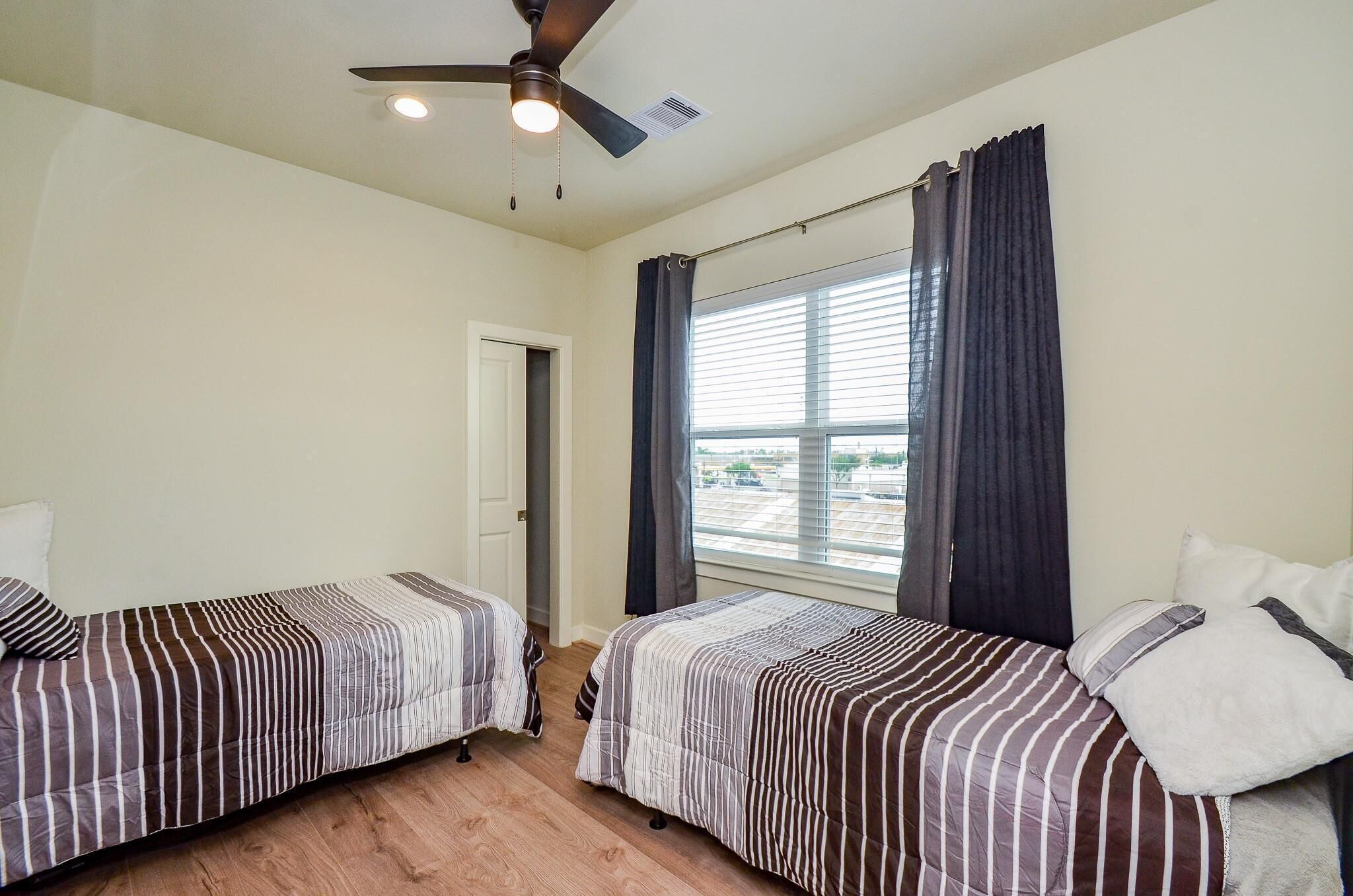 A bedroom with two single beds, a ceiling fan, a window with curtains, and a door.