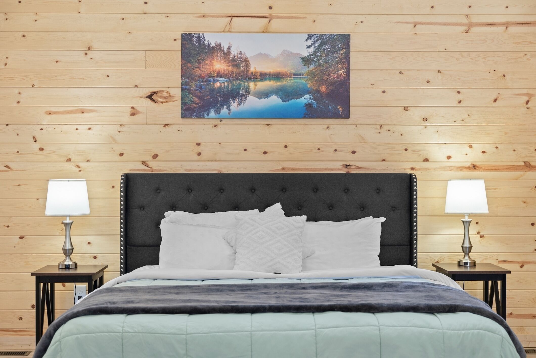 A bedroom with a wooden paneled wall, a bed with a dark headboard, two bedside lamps, and a framed picture above the bed.