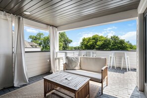 A covered outdoor seating area with a wooden table, a sofa, and a view of trees and buildings.