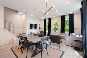A modern living room with a dining area, a staircase, and a large window with curtains.