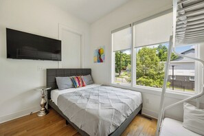 A bedroom with a bed, a TV mounted on the wall, a window with a view of trees, and a spiral staircase. - 16 Bed Private Gulch Home with Rooftop (Nashville)