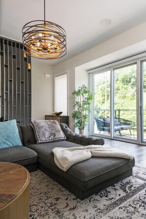 A modern living room with a sectional sofa, a wooden coffee table, and a large window with a view of greenery. - 19 Bed Home Near Downtown with Game room (Nashville)