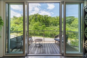 A balcony with a wooden deck, two chairs, a small round table, and a view of a green landscape.