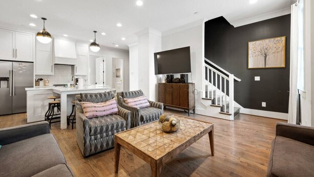 Un salon moderne avec une table basse en bois, deux fauteuils à motifs, un canapé et une cuisine dotée de meubles blancs. Un escalier mène à un mur sombre orné d’un tableau encadré.