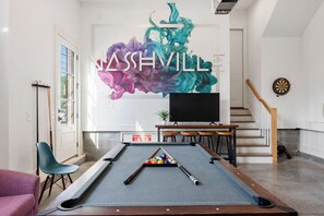 Una mesa de billar en una habitación con un mural, una televisión y un dardo.