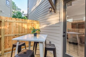 A patio with a table and two stools, a wooden fence, and a view into a house through a glass door.