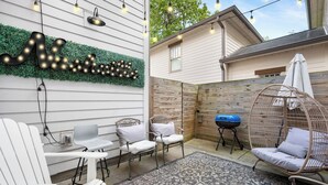 Un jardin avec une guirlande lumineuse, un barbecue, des chaises blanches et un fauteuil suspendu.