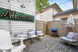 Un giardino sul retro con luci appese, un barbecue, sedie bianche e una poltrona amaca.