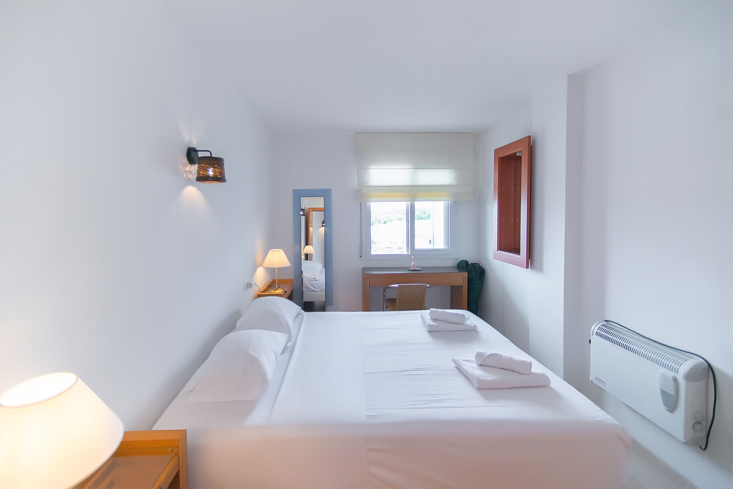 Une chambre d’hôtel comprenant un lit, une table de chevet, une lampe, un bureau, une chaise, une fenêtre et un radiateur.
