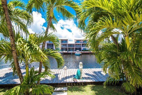 A wooden dock with a boat, palm trees, and a building with balconies by a body of water.