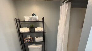 Une petite salle de bain comprenant des toilettes, une étagère avec des serviettes et des articles de toilette, un rideau de douche et un petit tapis.