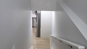 A narrow staircase with wooden steps and white walls, leading to a kitchen area with a refrigerator and cabinets.