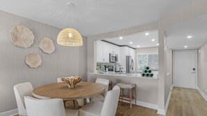 Een moderne keuken met witte kasten, een eethoek met een ronde houten tafel en witte stoelen, en een hanglamp.