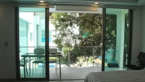 Habitación de hotel moderna con un amplio ventanal corredero que da a un balcón con vistas a un árbol y una piscina.