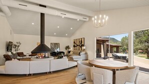 Tilava olohuone, jossa on suuri takka, ruokailutila pyöreän pöydän ympärillä sekä näkymä ulos.