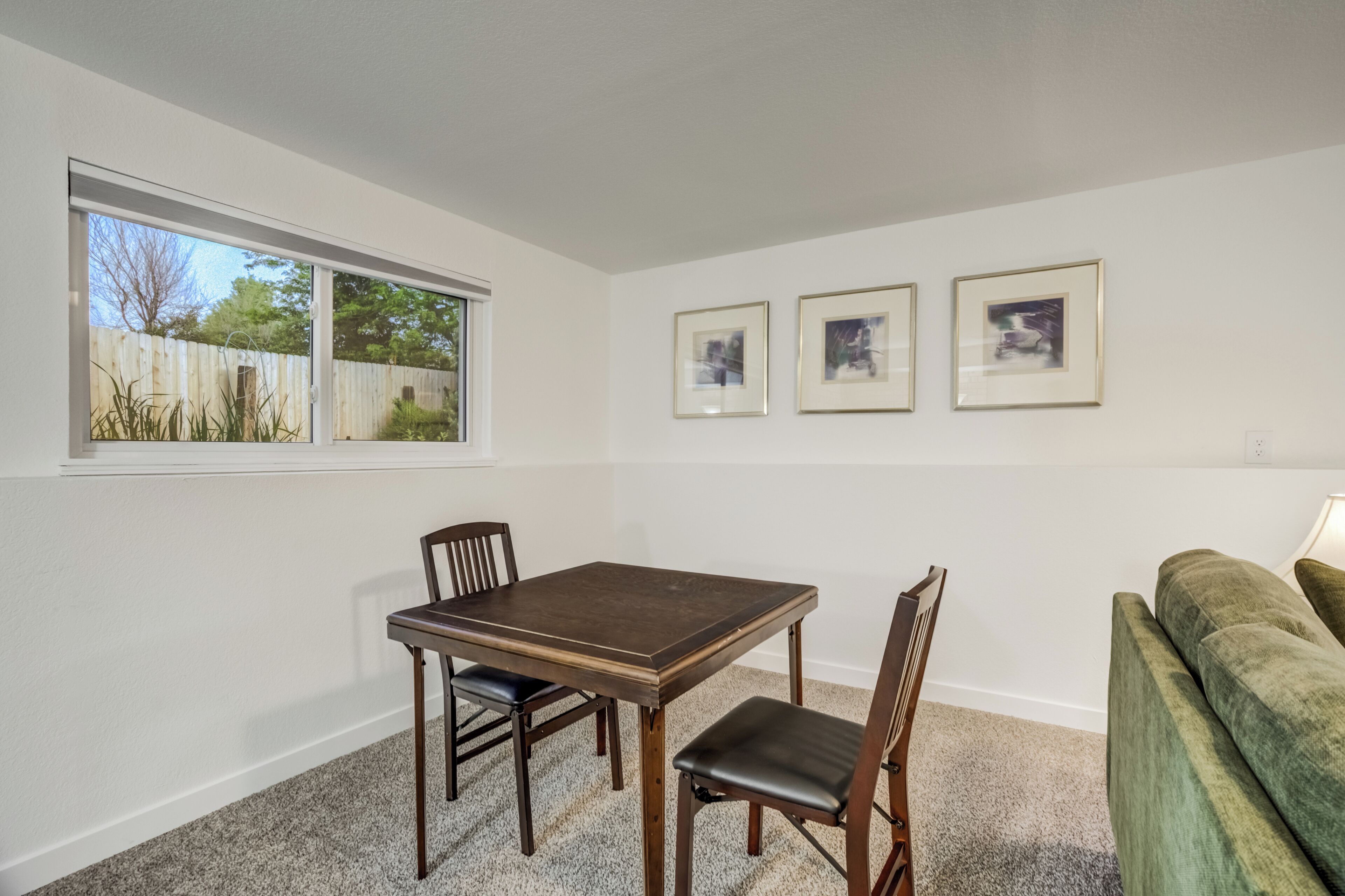 Un comedor con una mesa y sillas, una ventana con vista a una cerca de madera y árboles, y tres cuadros en la pared.