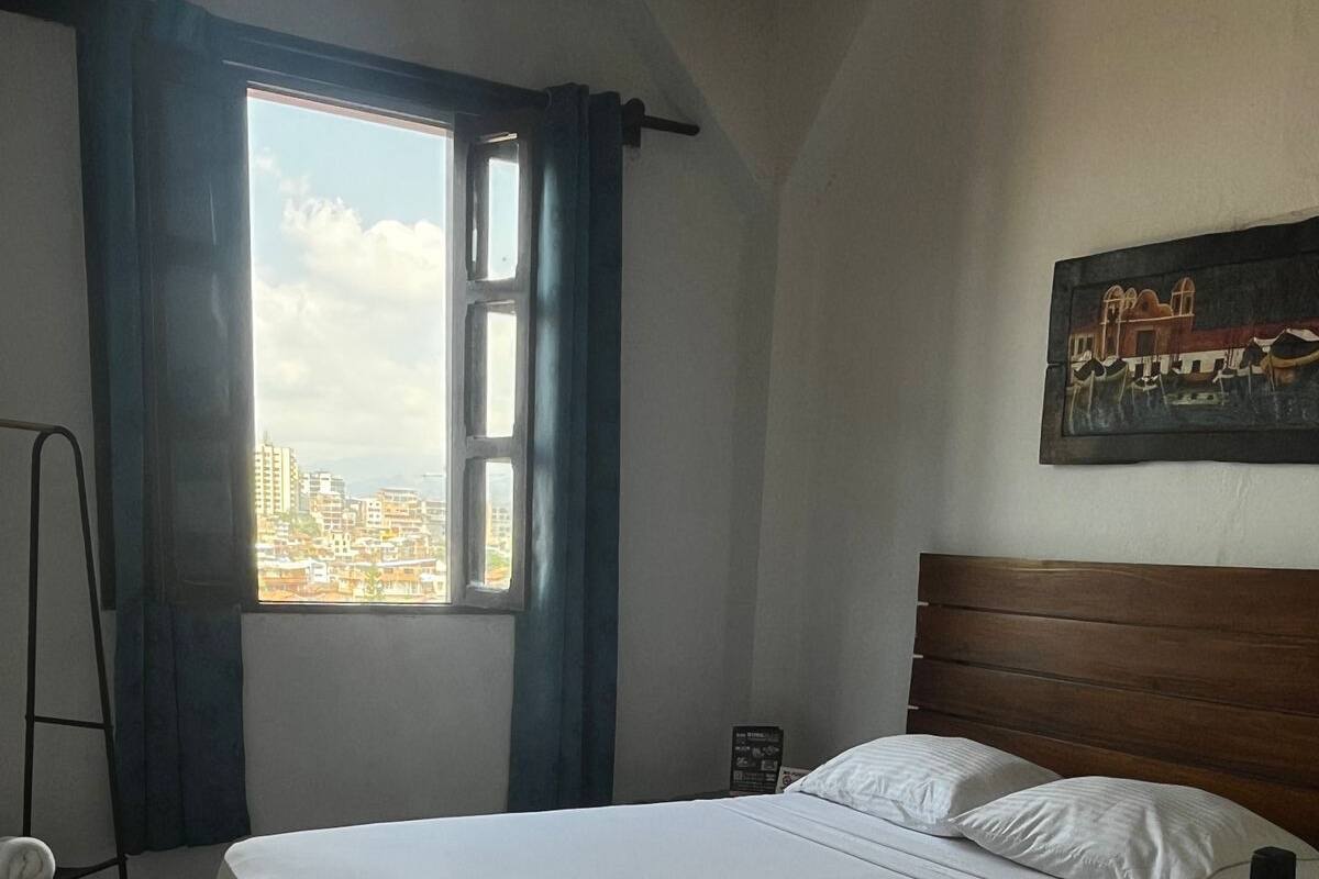 Ein Schlafzimmer mit Bett, einem Fenster mit Vorhängen, einem Sessel und einem Bild an der Wand.