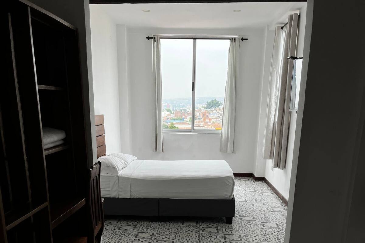 Ein Zimmer mit einem Bett, einem Sessel, einem Kleiderschrank und einem Fenster mit Vorhängen.