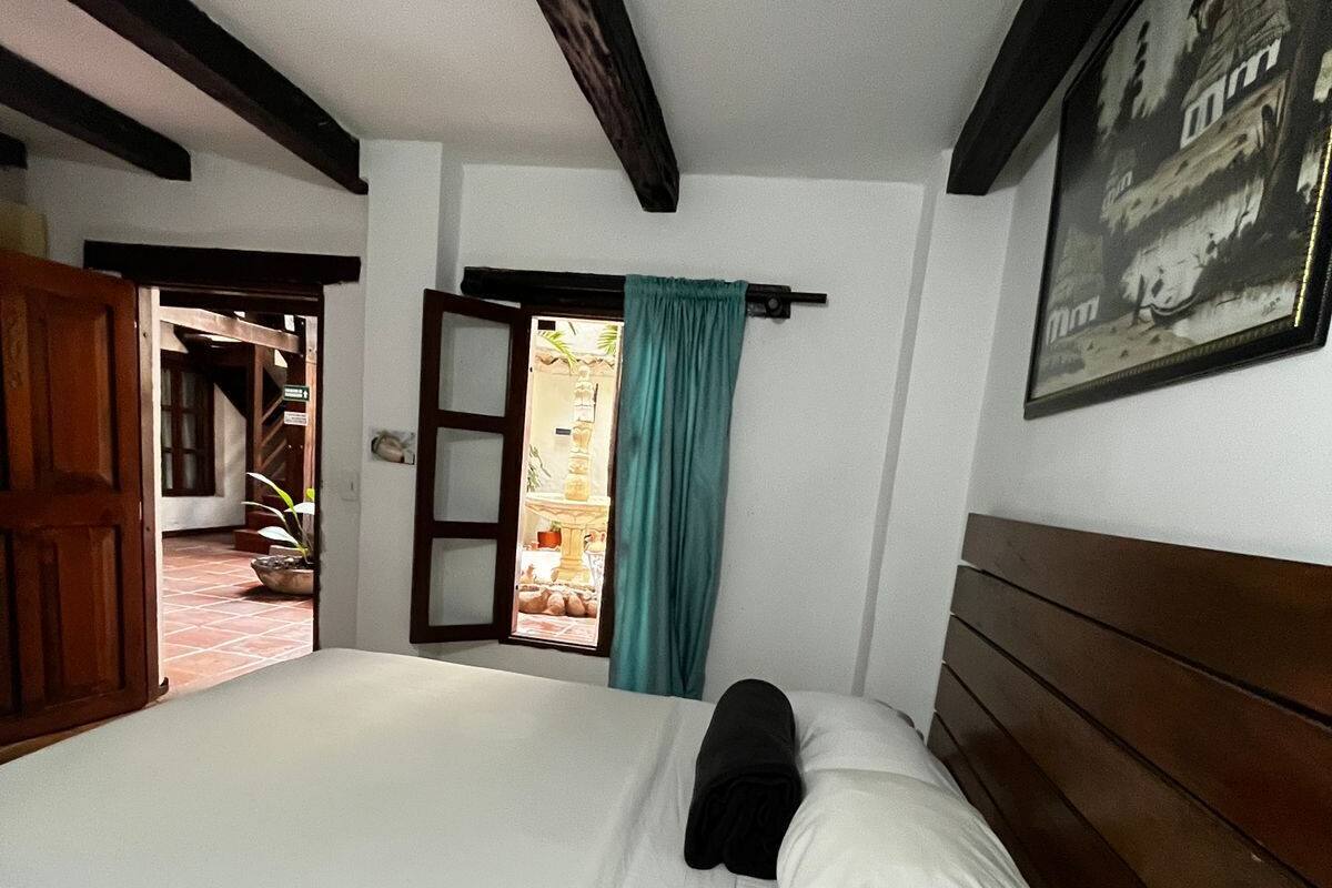 Ein Zimmer mit einem Bett, einer Holztür, einem mit Vorhängen versehenen Fenster und einem gerahmten Bild an der Wand.