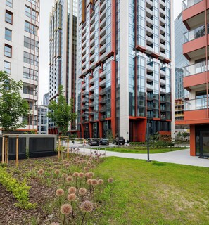 A modern urban residential building with multiple floors, large windows, and a well-maintained garden area with shrubs and flowers.