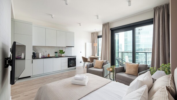A modern hotel room with a large bed, a sofa, a small table, and a balcony with a view of the city.