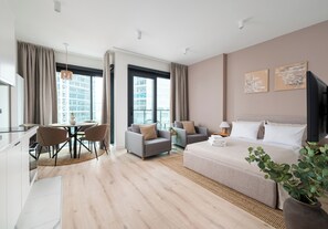 A modern hotel room with a large bed, a dining area with chairs, a sofa, and a view of the city through large windows.