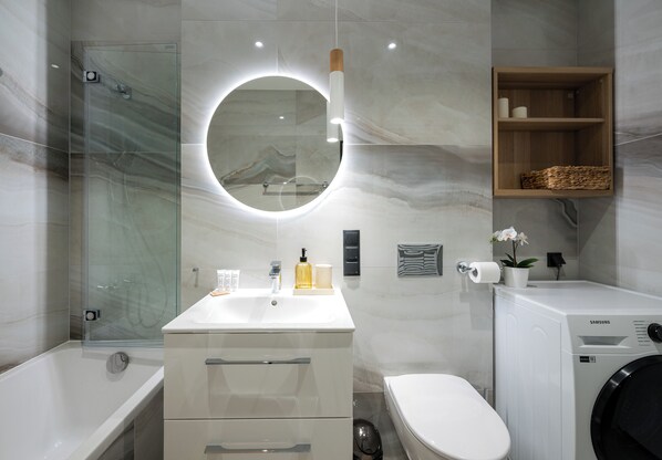 A modern bathroom with a glass-enclosed shower, a round mirror with a light, a white sink, a toilet, and a washing machine.