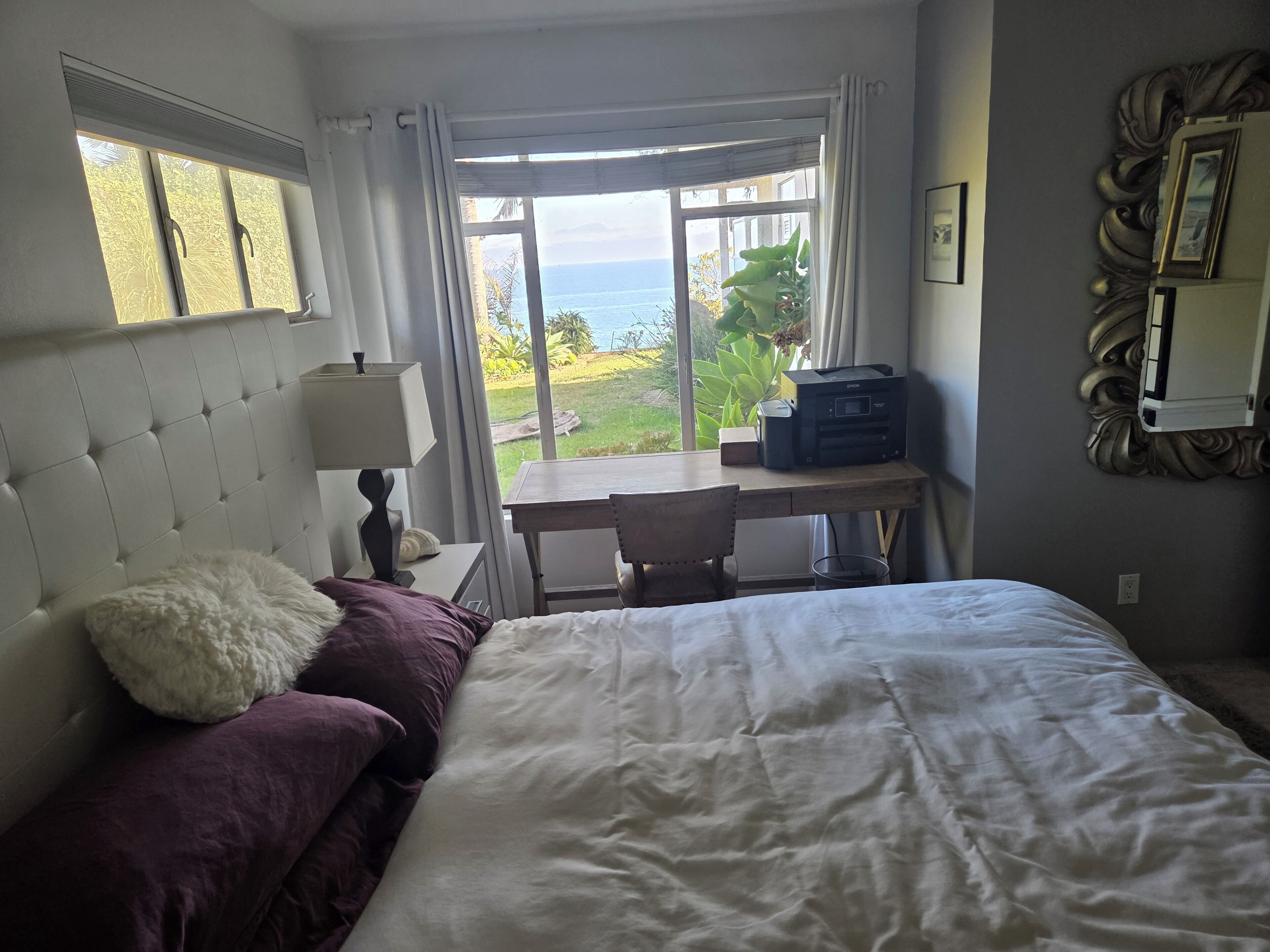 A hotel room with a bed, a desk, a lamp, a printer, and a view of the sea.