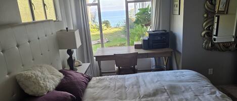 A hotel room with a bed, a desk, a lamp, a printer, and a view of the sea.