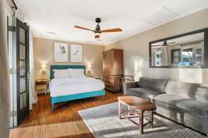 Un dormitorio con cama, sofá gris, armario de madera, ventilador de techo y un espejo grande.