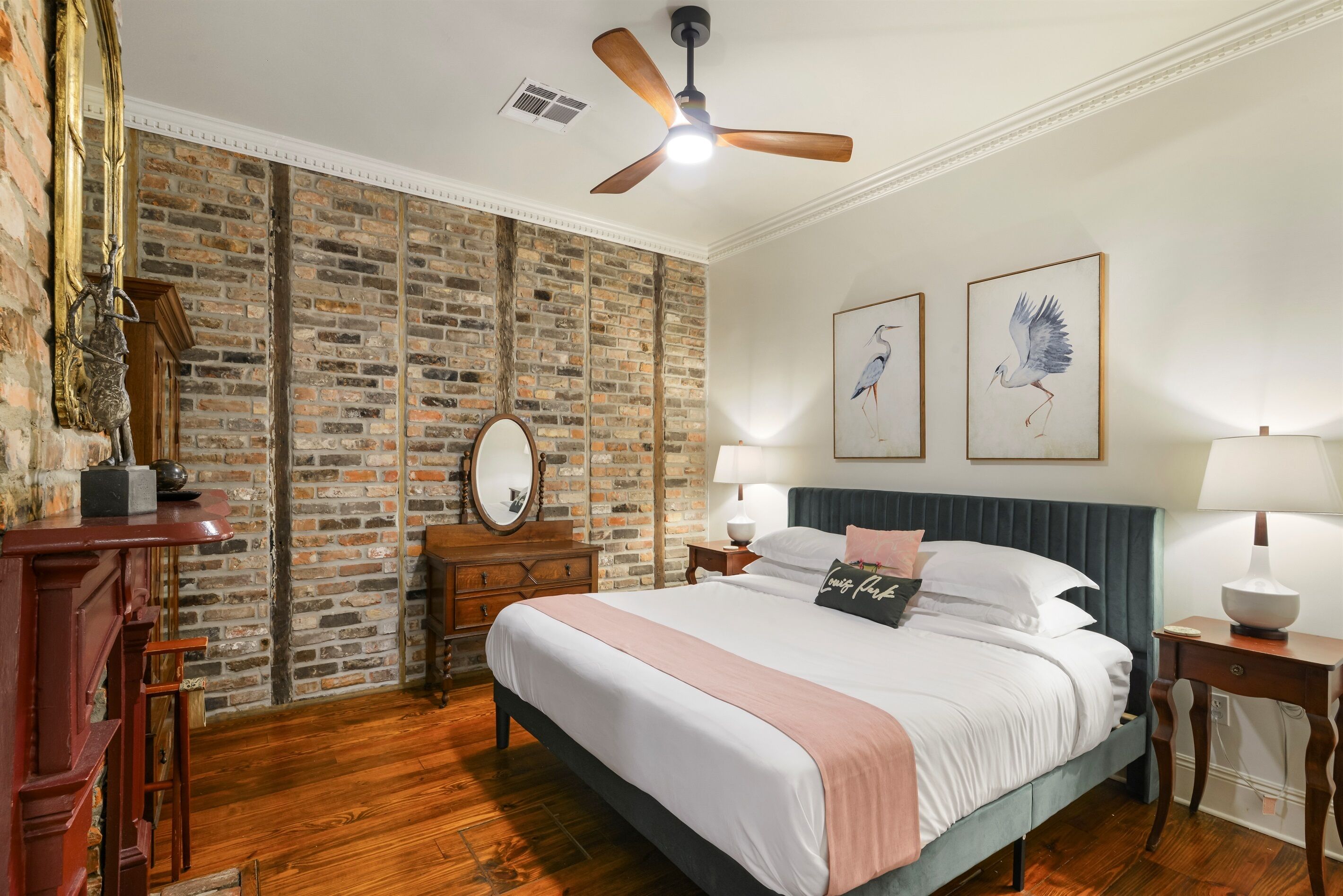 A bedroom with a brick wall, a bed with a blue headboard, two wall-mounted lamps, and a ceiling fan.