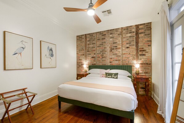 A bedroom with a brick-patterned wall, a green upholstered headboard, a wooden bed frame, a wooden bench, and two wall-mounted lamps.