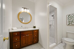 Un baño con tocador de madera, un espejo redondo con luces, inodoro blanco y zona de ducha.