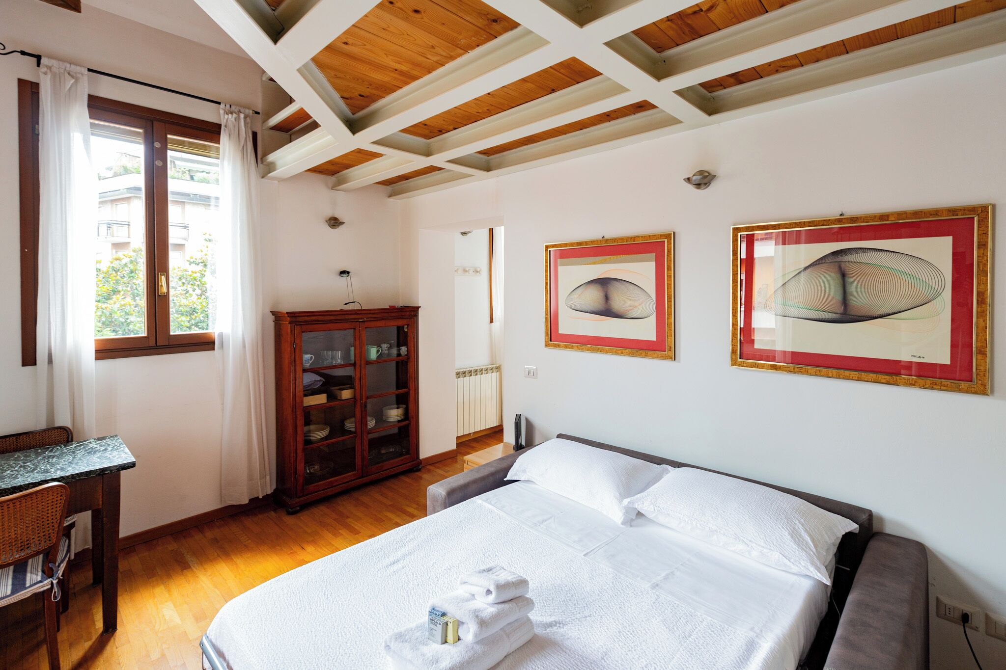 A hotel room with a bed, a desk, a cabinet with dishes, and two framed artworks on the wall.