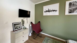 Ein Wohnzimmer mit Flachbildfernseher an der Wand, einem roten Sessel, einer weißen Kommode mit einem gerahmten Bild und zwei gerahmten Stadtansichten an der Wand.