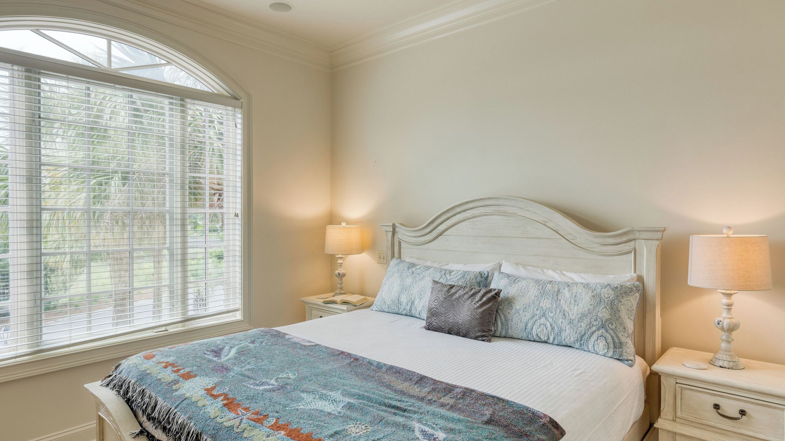 A bedroom with a large window, a bed with patterned bedding, two bedside lamps, and a nightstand.