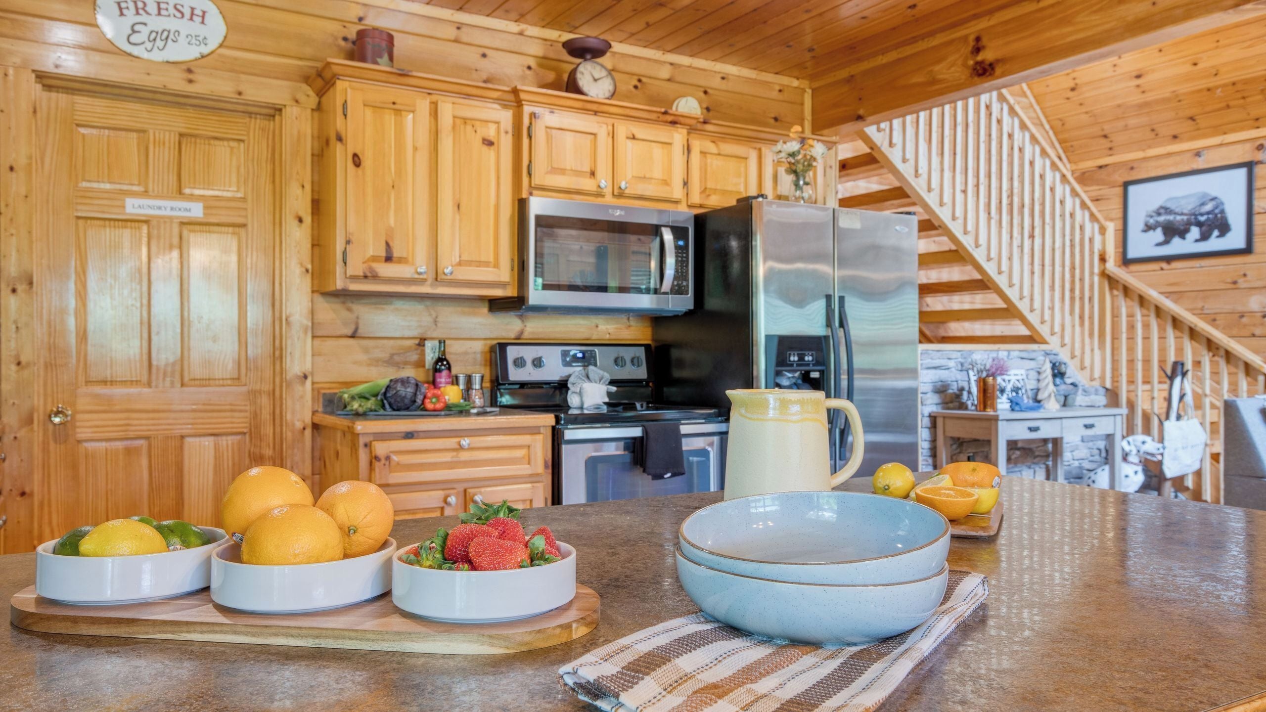 A kitchen with wooden cabinets, a microwave, a refrigerator, and a stove. There are bowls with fruits on the counter.