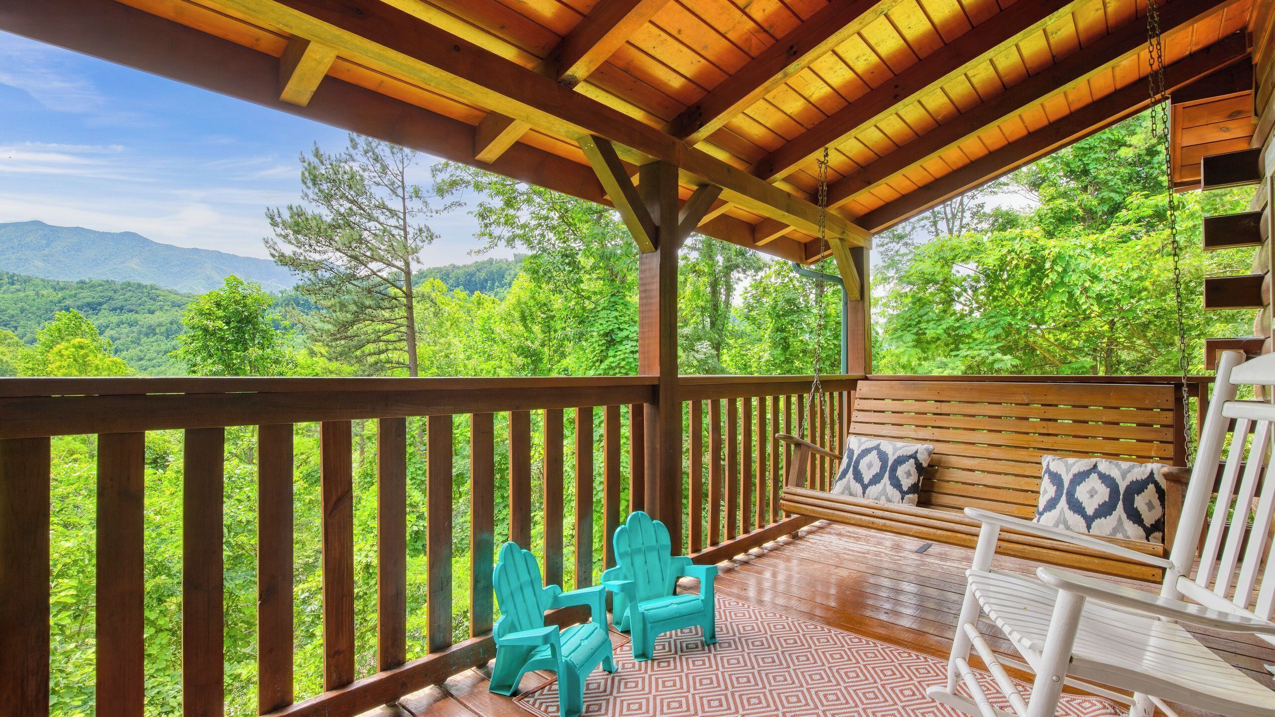 Une terrasse en bois offrant une vue sur les montagnes, deux fauteuils Adirondack turquoise et une chaise à bascule blanche avec des coussins à motifs.