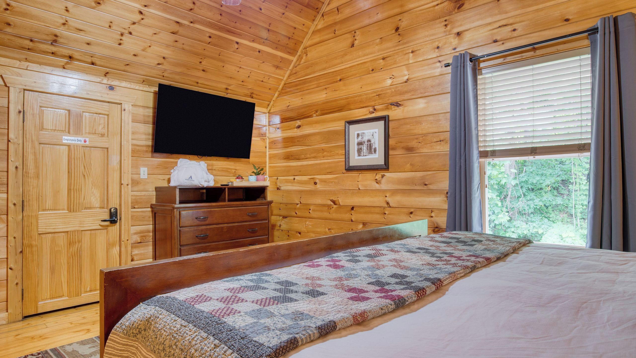 Een houten kamer met een bed, een dressoir, een televisie en een raam met jaloezieën.