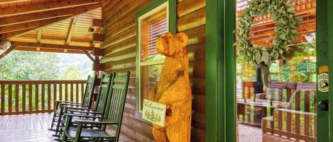 Una mecedora de madera en un porche con un letrero de 'Welcome', una escultura de oso de madera y un adorno en una ventana.
