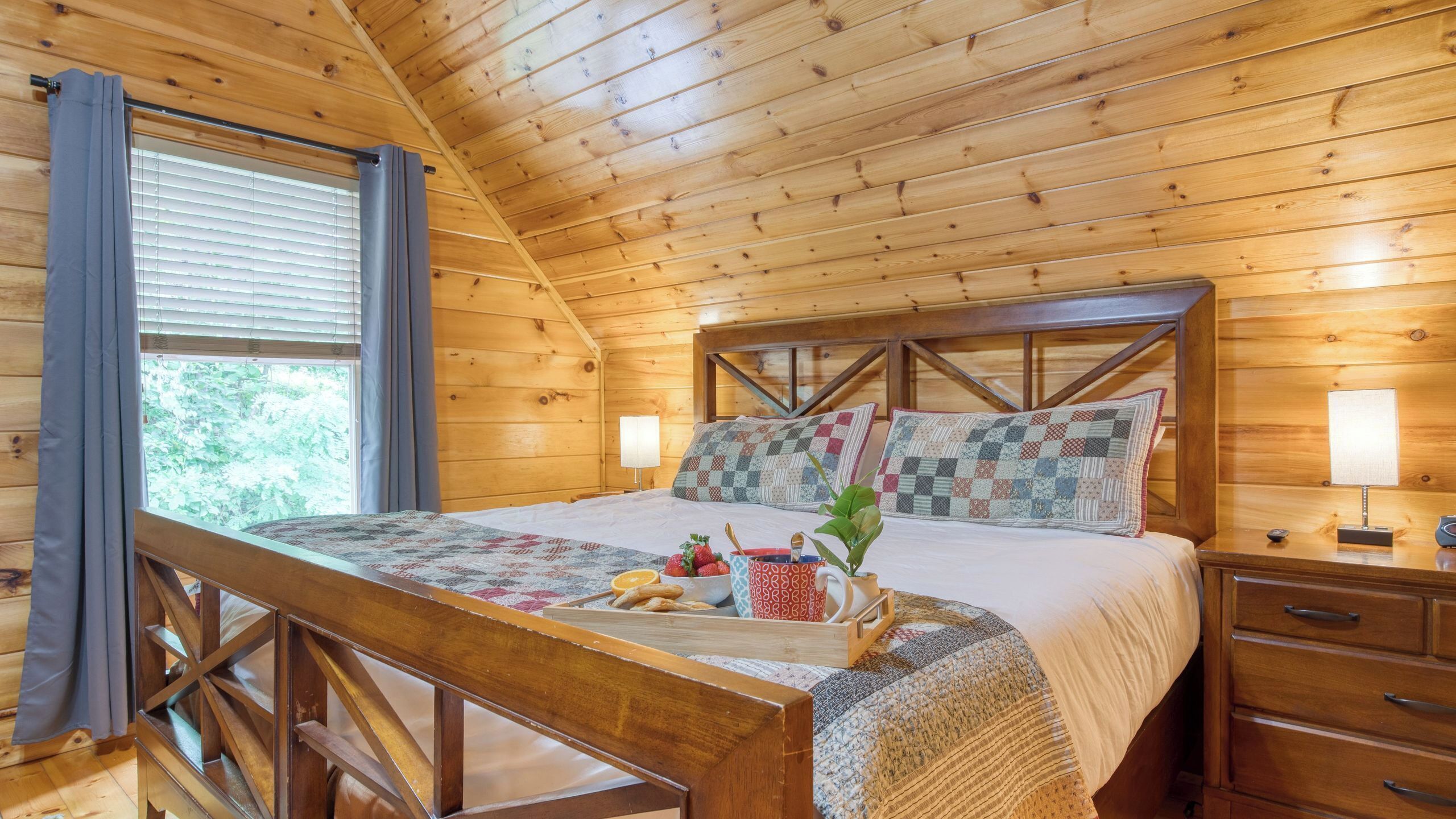 Ein Schlafzimmer einer Holzhütte mit Bett, Nachttischen, einem Fenster mit Vorhängen und Blick auf Grünland.