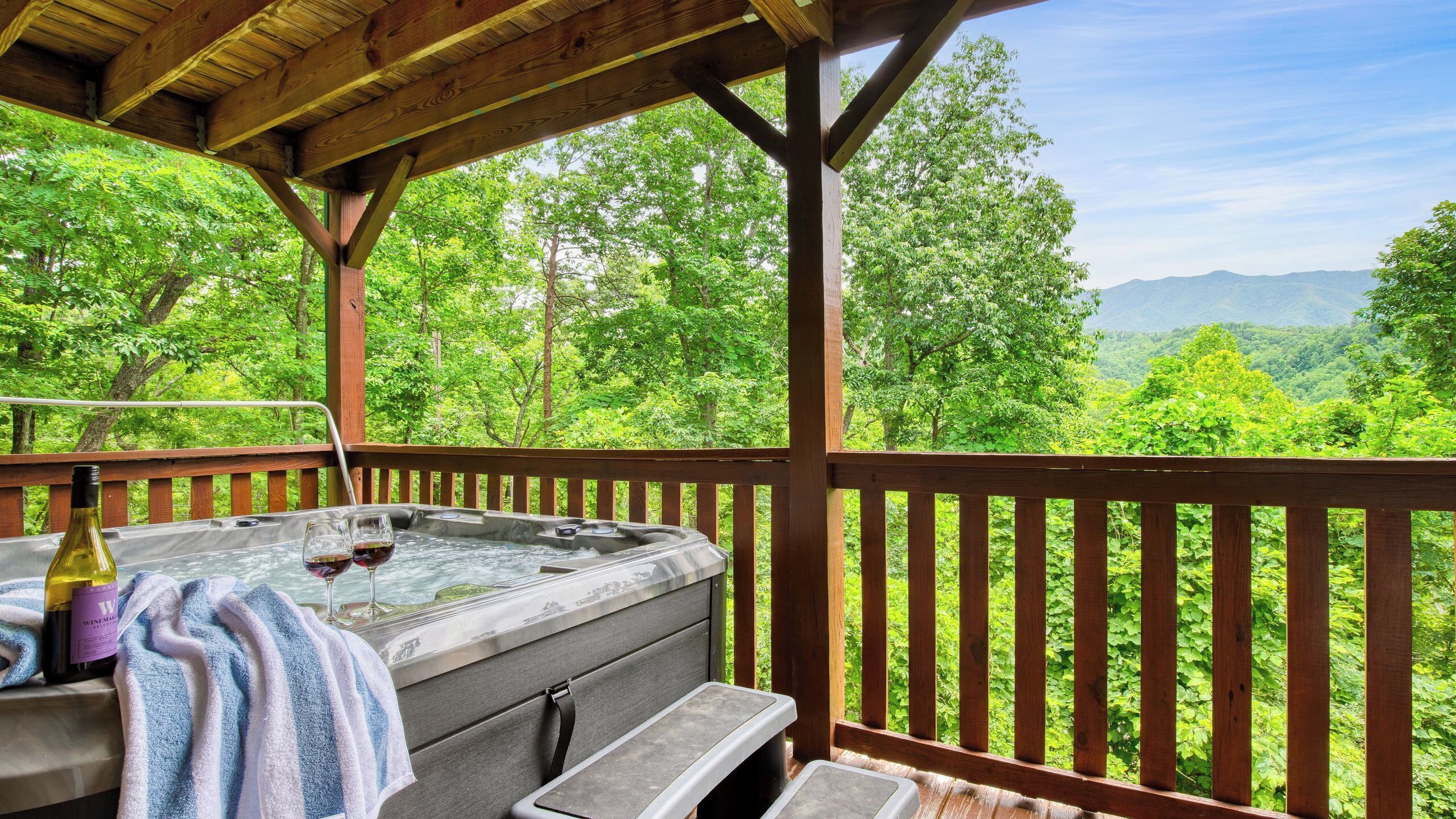 A hot tub with a towel and wine bottle, wooden deck, and a view of lush greenery and mountains.