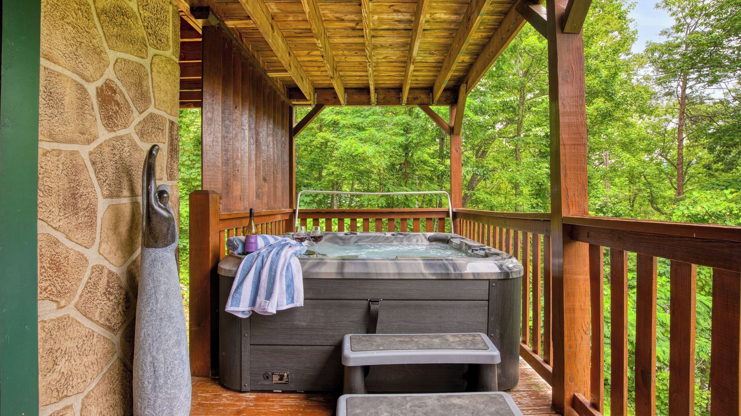 A hot tub under a wooden pergola with stone walls, surrounded by lush greenery.