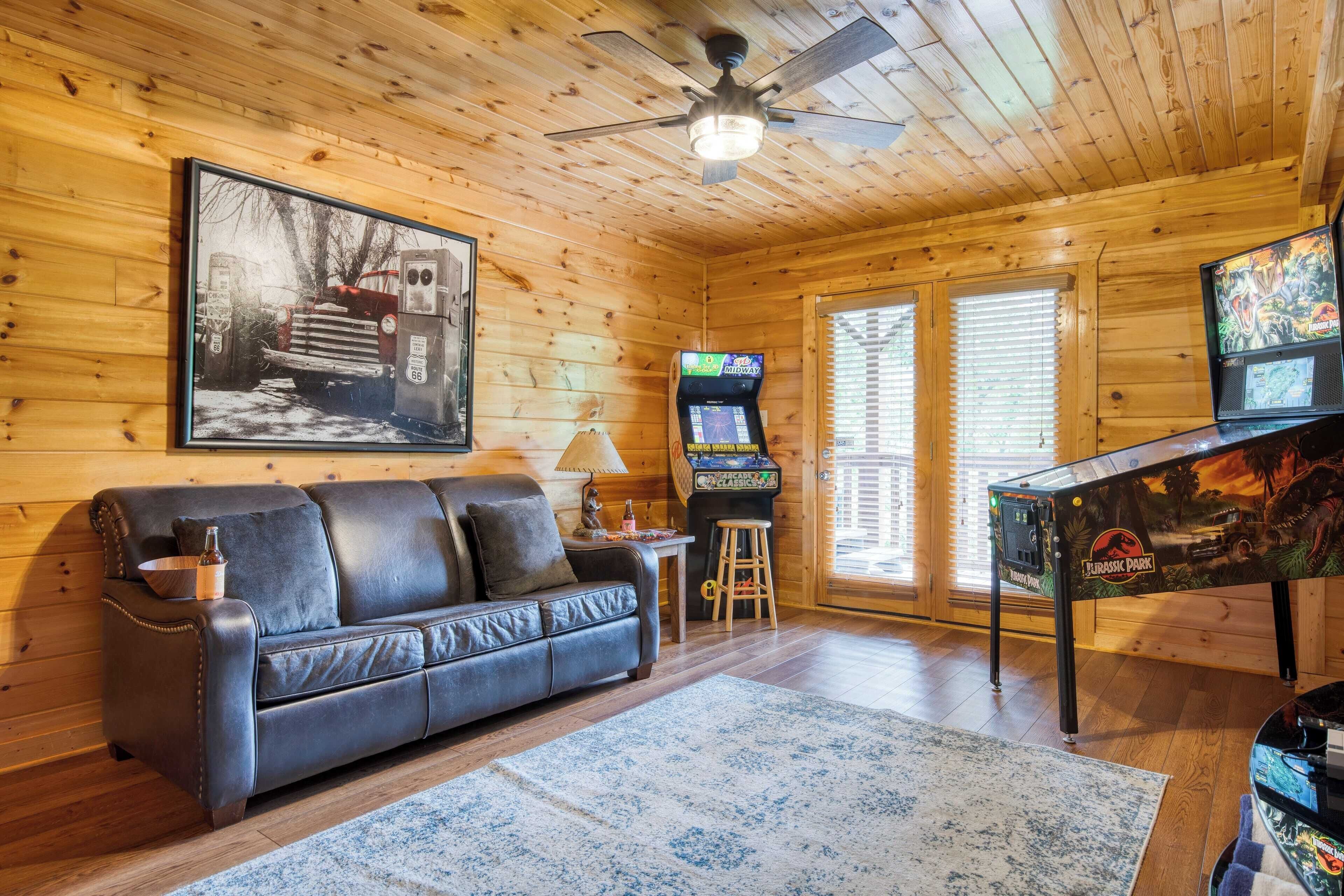 A cozy cabin interior with a wooden ceiling, a black leather sofa, a gaming table, and a large framed picture on the wall.