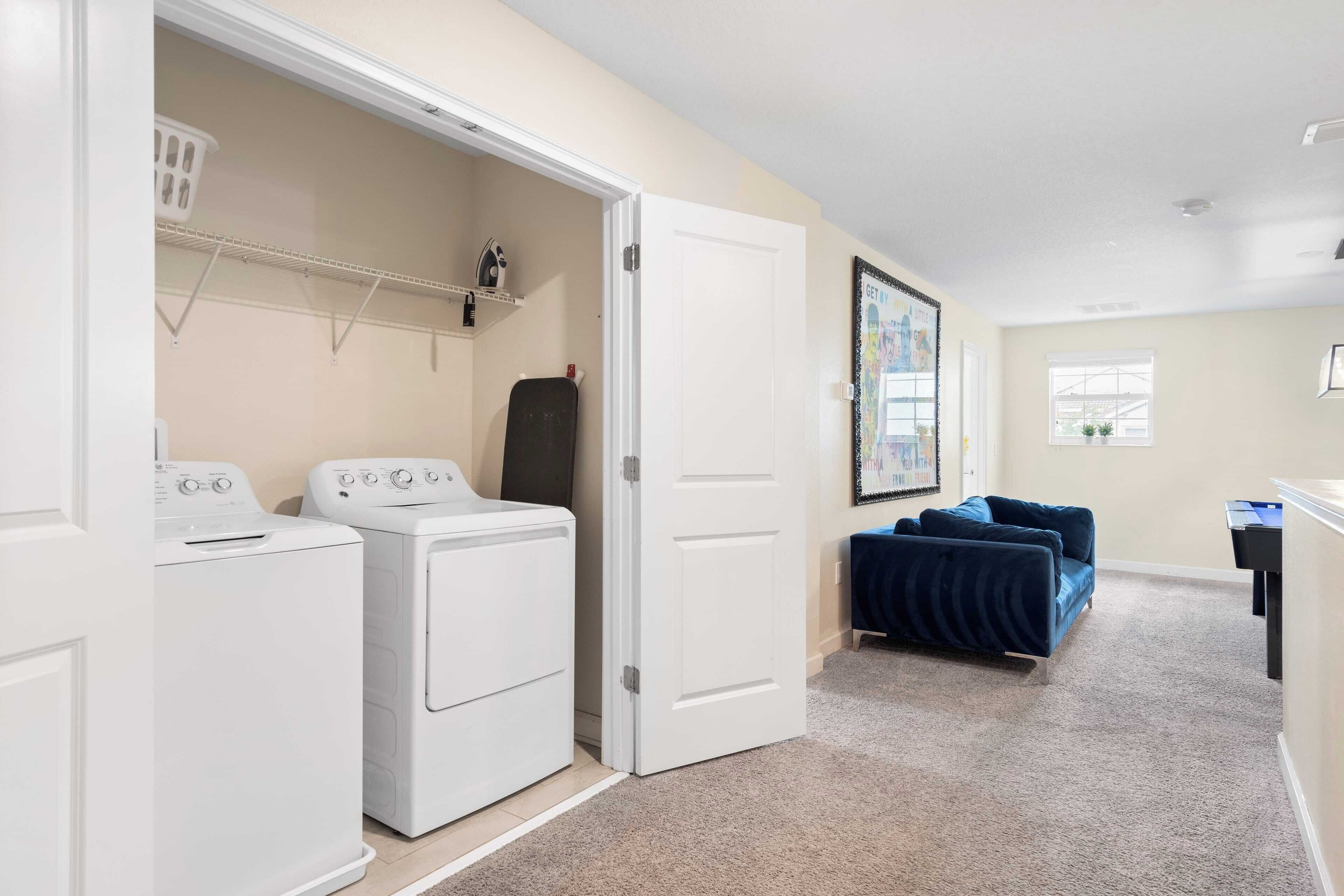 A laundry room with two white washing machines, a drying rack, and a door leading to a living area with a blue couch and a small table.