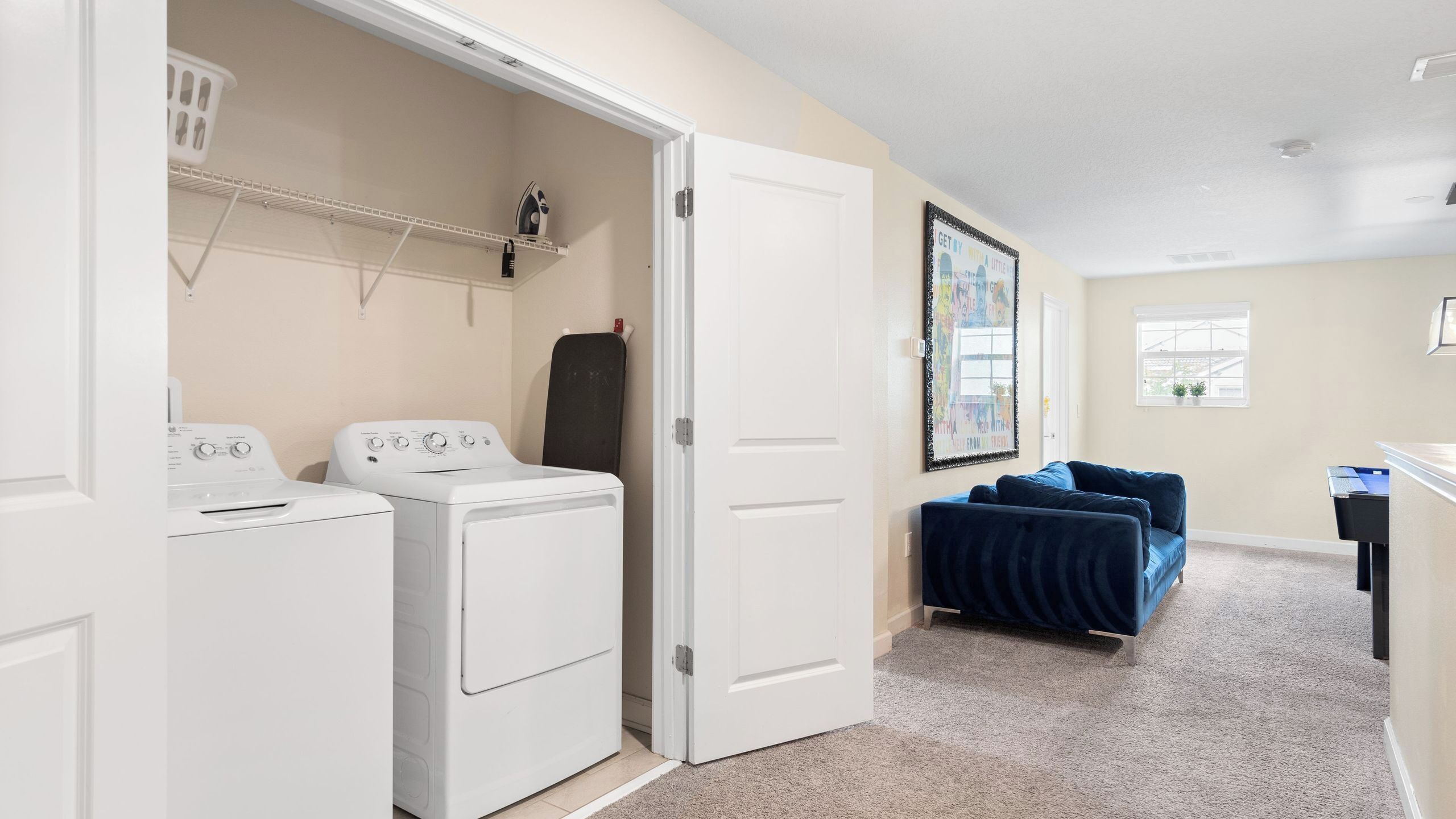 A laundry room with two washers and dryers, a door leading to another room, a blue couch, and a framed picture on the wall.