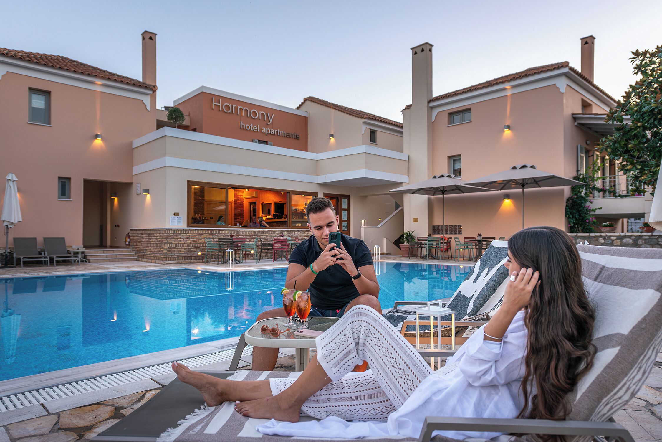 A poolside area with lounge chairs, a bar, and a building labeled "Harmony Hotel Apartments."