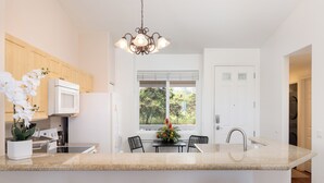 A modern kitchen with a granite countertop, wooden cabinets, a central island with bar stools, a chandelier, and a view of the outdoors through a window.