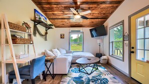 Una habitación acogedora con ventilador de techo, una cama, un escritorio con silla, televisión y ventana con vistas a follaje.
