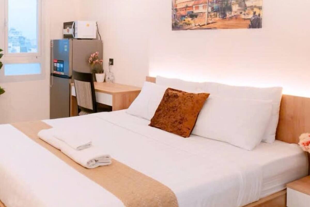 A neatly made bed with white linens and a brown pillow, a bedside table with a lamp, and a small desk with a computer and a plant.